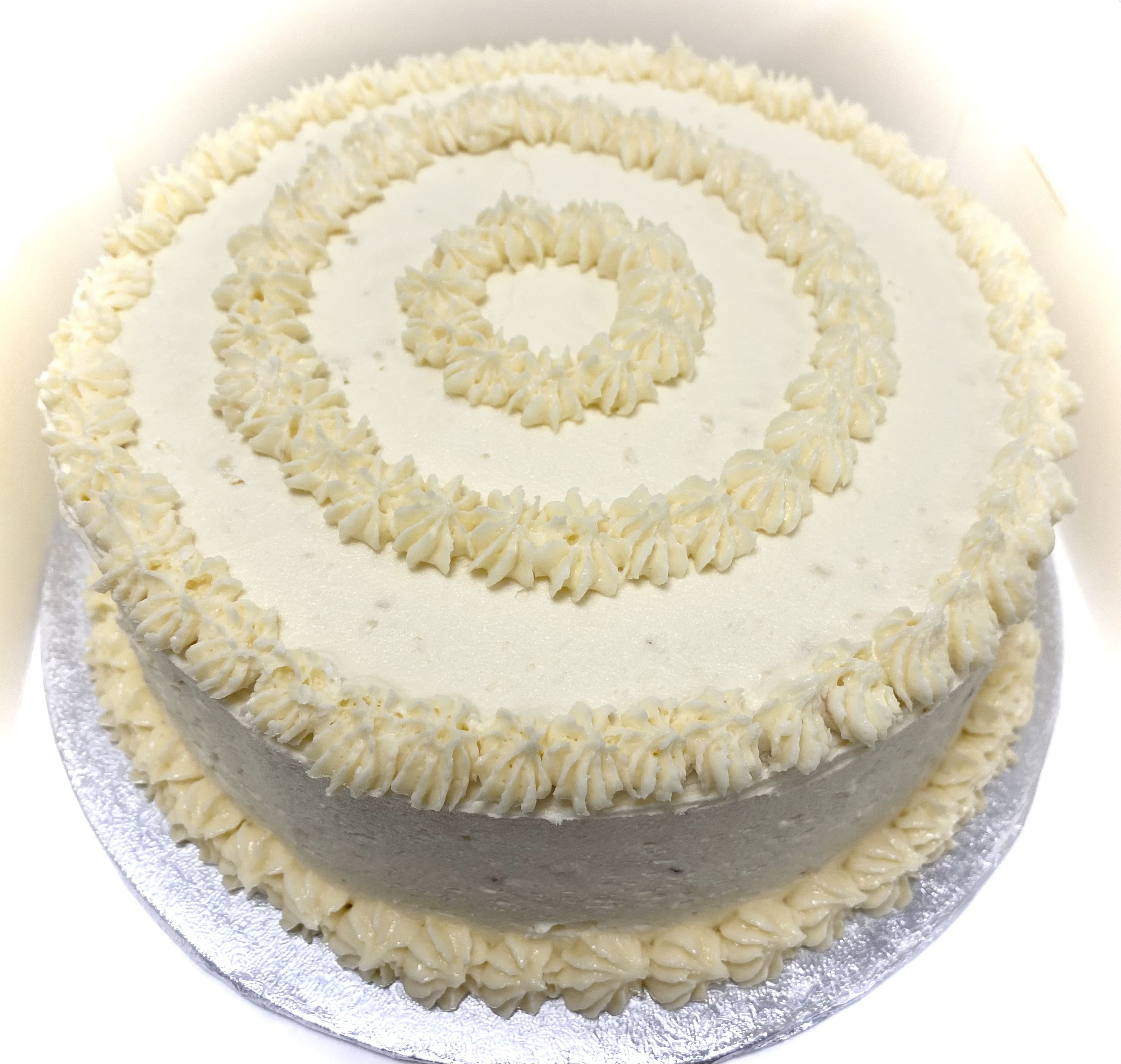 A round, frosted Vanilla Gateaux with white icing, adorned with concentric circles of piped frosting on the top and evenly spaced piped frosting along the top edge. This classic cake sits elegantly on a reflective silver cake board, making it the go-to dessert for any celebration.
